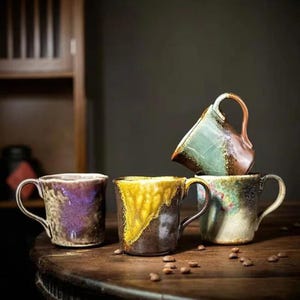 May include: Four ceramic mugs with unique, textured finishes. The mugs feature a variety of colours, including purple, yellow, green, and brown. Each mug has a curved handle. The mugs are arranged on a wooden surface with scattered coffee beans.