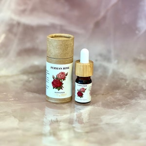 Op de afbeelding: Een kleine bruine kartonnen buis met een wit label waarop "Persian Rose" staat en een kleine rode roos illustratie. De buis staat naast een klein glazen flesje met een bamboe druppelaar en een wit label waarop "HANNANEH" staat en een kleine rode roos illustratie.
