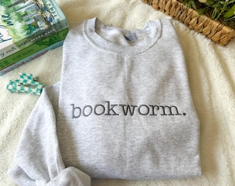 May include: Gray sweatshirt with the text "bookworm." embroidered on the front. The sweatshirt is laying on a white fuzzy surface.