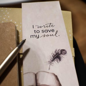 Può includere: Un segnalibro con una piuma e un'illustrazione di un libro aperto. Il testo sul segnalibro recita "Scrivo per salvare la mia anima."