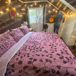 Könnte beinhalten: Ein Bett mit einem rosafarbenen Bettbezug und passenden Kissenbezügen mit einem dunkelvioletten botanischen Blattmuster. Das Bett steht in einem Raum mit dunklen Wänden, Lichterketten und zwei lebensgroßen Pappaufstellern von Personen.