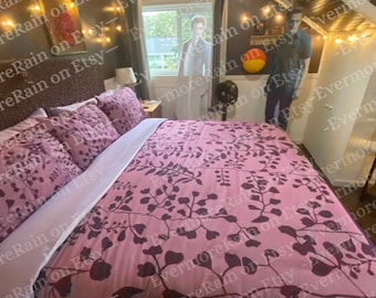 Puede incluir: Una cama con funda nórdica rosa y fundas de almohada a juego con un estampado botánico de hojas en color morado oscuro. La cama se encuentra en una habitación con paredes oscuras, luces de cadena y dos figuras de cartón de personas.