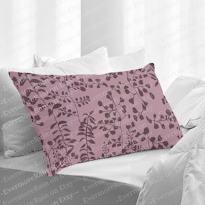 May include: A pink pillow with a floral pattern featuring dark brown leaves and branches. The pillow is on a white bed with a white blanket.