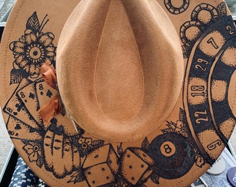 May include: A brown felt fedora hat with a black ink design of playing cards, dice, a roulette wheel, and a pool ball. The hat is surrounded by playing cards and chips.