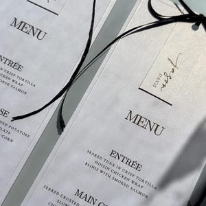May include: White menu cards with black text and a black ribbon. The menu lists dishes such as seared tuna, chicken, and dessert options. The top of the card has the word "MENU" and a signature.