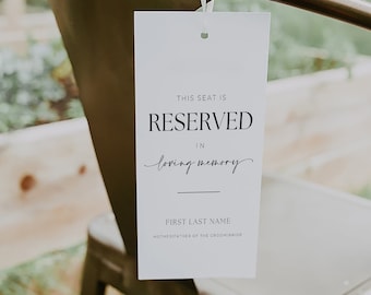 May include: White card with black text that reads "This seat is reserved in loving memory" with a line for a name and the words "Mother/Father of the Groom/Bride". The card is tied to a metal chair with a white string.