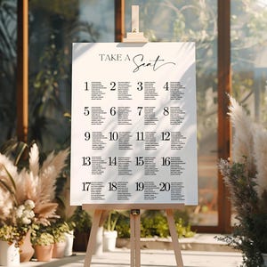 May include: A white seating plan with the words "TAKE A Seat" in elegant script. The chart is displayed on a wooden easel and lists names under numbers 1-20. The background features plants and natural light.