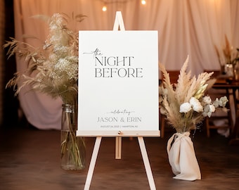 May include: White sign with black text on an easel, reading "The Night Before celebrating Jason & Erin August 24, 2023 | Hampton, NJ". The sign is in front of a white backdrop with string lights and dried flowers.