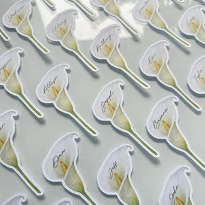 May include: A collection of die-cut paper place cards shaped like white calla lilies with yellow centers. Each individual flower features a unique name printed in black cursive script across the petal, arranged in a repeating pattern.