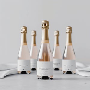 Könnte beinhalten: Fünf Champagnerflaschen mit goldfarbenen Verschlüssen und Etiketten. Die Flaschen sind mit einer hellrosa Flüssigkeit gefüllt. Auf den Etiketten steht "Bridesmaid" und "Maid of Honor" mit den Worten "Prosecco Rose" und einem Datum. Die Flaschen stehen auf einer weißen Oberfläche.