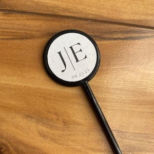 May include: A black cocktail stirrer with a circular white top. The top features the initials "J|E" and the date "08.25.23" in a simple, elegant font. The stirrer is set against a wooden background.
