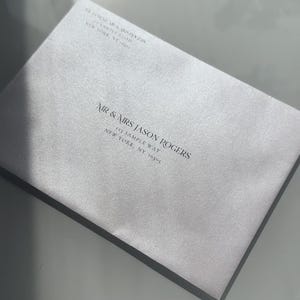 May include: A light gray envelope with the names "NIR & MRS JASON ROGERS" and address details printed in dark gray. The envelope is on a reflective surface.