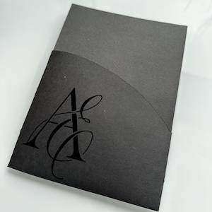 Könnte beinhalten: Ein schwarzer Einladungsumschlag mit gebogenem oberem Klappdeckel. Auf der Vorderseite befindet sich ein stilisiertes, geprägtes Monogramm der Buchstaben "A" und "C" in einer fließenden Schrift. Der Umschlag besteht aus dickem, mattem Papier.
