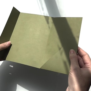 Pode incluir: Um cartão verde-oliva dobrado, segurado entre duas mãos. O cartão é feito de papel texturizado e tem um brilho sutil. O fundo é branco com sombras.