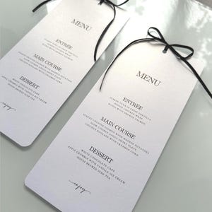 Op de afbeelding: Twee witte menukaarten met zwarte linten. Elke kaart toont het woord "MENU" en een lijst met gerechten, waaronder Voorgerecht, Hoofdgerecht en Dessert. De menu's liggen op een reflecterend oppervlak.