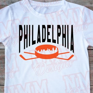 May include: White t-shirt featuring a black graphic of the word "PHILADELPHIA" with a hockey stick and puck design underneath. The puck depicts a silhouette of the Philadelphia skyline.