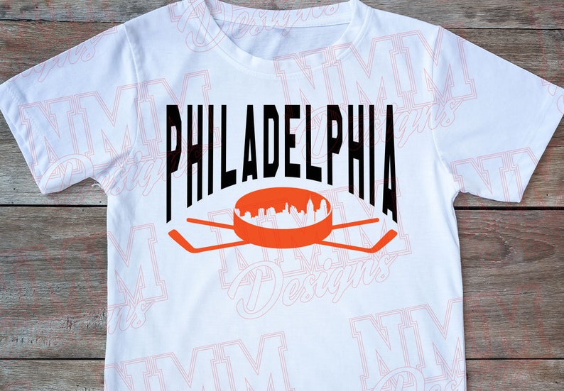 May include: White t-shirt featuring a black graphic of the word "PHILADELPHIA" with a hockey stick and puck design underneath. The puck depicts a silhouette of the Philadelphia skyline.