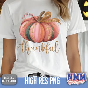 May include: White t-shirt with a watercolour pumpkin design in pink, orange, and green, with gold glitter. The word "thankful" is written in gold script below the pumpkin. A digital download graphic.