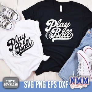May include: Two white t-shirts with black text that says "Play Ball" and a baseball graphic. The shirts are on a wooden surface with a pair of jeans and sneakers.