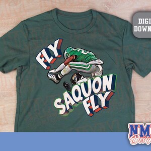 K&ouml;nnte beinhalten: Gr&uuml;nes T-Shirt mit einem Footballspieler in gr&uuml;n-wei&szlig;er Uniform und den Worten "FLY SAQUON FLY" in 3D-Design. Das Shirt hat auch eine "DIGITAL DOWNLOAD" Grafik und das "NMM Designs" Logo.