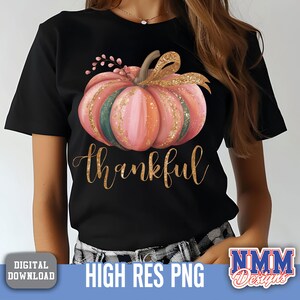 May include: Black t-shirt with a watercolour pumpkin design in pink, orange, and green, with gold glitter accents. The word "thankful" is written in gold script below the pumpkin. A fall-themed graphic tee.