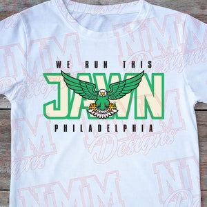 Pu&ograve; includere: Maglietta bianca con una grafica verde e crema di un'aquila con il testo "WE RUN THIS JAWN PHILADELPHIA".