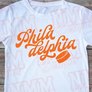 May include: White t-shirt with an orange graphic that says "Phila delphia" with a hockey puck underneath.