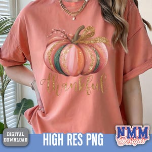 May include: A coral t-shirt with a pumpkin design in watercolour, featuring pink, orange, and green sections, with gold glitter and a gold bow. The word "Thankful" is written in gold script below the pumpkin. The shirt has rolled-up sleeves.