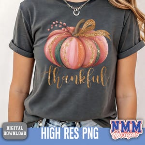 May include: Dark grey t-shirt with a watercolour pumpkin design in pink, green, and gold. The word "thankful" is written in gold script below the pumpkin. A digital download graphic.