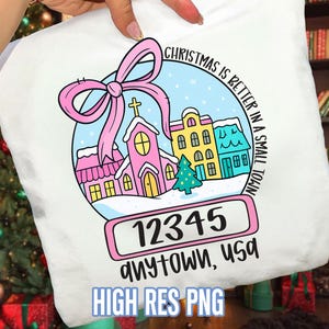 May include: White tote bag featuring a colorful illustration of a small town in a snow globe. The design includes a pink church, houses, and a Christmas tree, with the text "Christmas is better in a small town". The town's zip code is 12345.
