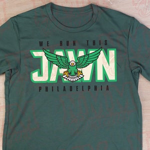 Pu&ograve; includere: Una maglietta verde con una grafica bianca di un'aquila calva con il testo "WE RUN THIS JAWN PHILADELPHIA" stampato sul davanti.