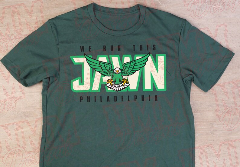 Pu&ograve; includere: Una maglietta verde con una grafica bianca di un'aquila calva con il testo "WE RUN THIS JAWN PHILADELPHIA" stampato sul davanti.