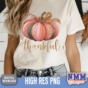 May include: Cream-coloured t-shirt with a watercolour pumpkin design in pink, orange, and green, with a gold glitter bow and the word "thankful" in gold script. Digital download.