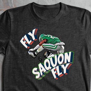 K&ouml;nnte beinhalten: Dunkelgraues T-Shirt mit einem Footballspieler in Gr&uuml;n und Wei&szlig;, der &uuml;ber einen anderen springt. Das Shirt zeigt die W&ouml;rter "FLY" und "SAQUON FLY" in einem 3D-Design.