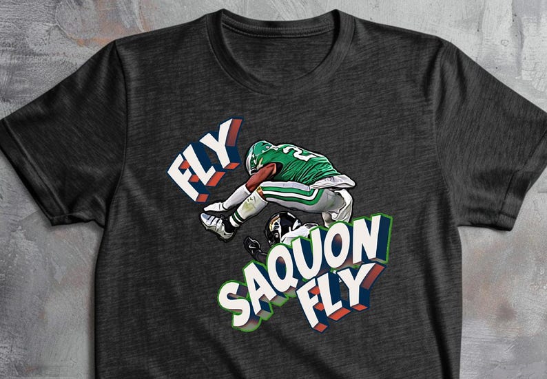 K&ouml;nnte beinhalten: Dunkelgraues T-Shirt mit einem Footballspieler in Gr&uuml;n und Wei&szlig;, der &uuml;ber einen anderen springt. Das Shirt zeigt die W&ouml;rter "FLY" und "SAQUON FLY" in einem 3D-Design.