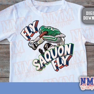 K&ouml;nnte beinhalten: Wei&szlig;es T-Shirt mit einer Grafik eines Footballspielers in gr&uuml;n-wei&szlig;er Uniform und den Worten "FLY SAQUON FLY" in 3D-Schrift. Das Design enth&auml;lt ein "DIGITAL DOWNLOAD"-Etikett.