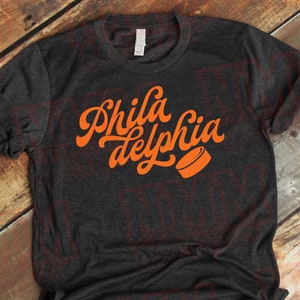 May include: A dark gray t-shirt with an orange graphic that says "Phila delphia" in a retro font with a hockey puck.