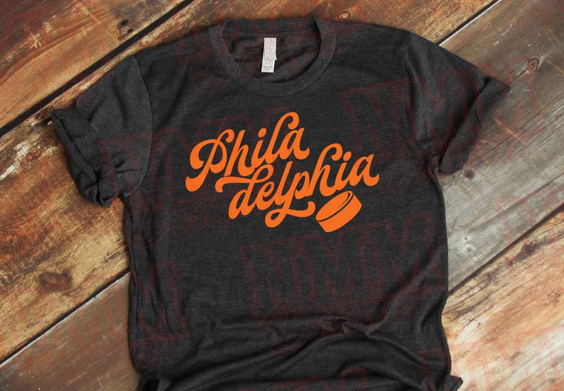 May include: A dark gray t-shirt with an orange graphic that says "Phila delphia" in a retro font with a hockey puck.