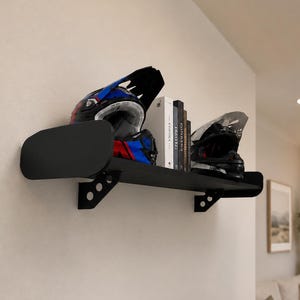May include: A black metal wall-mounted shelf displaying two motorcycle helmets and books. The helmets feature black, blue, and red designs. The shelf is fixed to a light-toned wall.