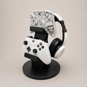Op de afbeelding: Een zwarte gamingstandaard met een witte en doorzichtige Xbox-controller en een wit-zwarte headset. De standaard heeft een cirkelvormige basis en een verticale ondersteuningsstructuur. De controllers hebben gekleurde knoppen.