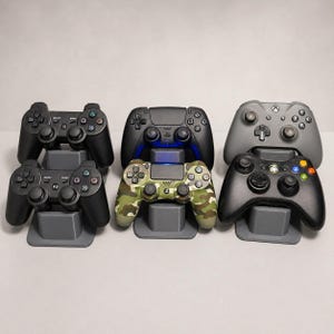 Può includere: Sei controller per videogiochi disposti in tre coppie su supporti in plastica grigia. La collezione comprende due controller neri, un controller nero a tinta unita, un controller con motivo mimetico, un controller grigio e un controller nero con pulsanti colorati.