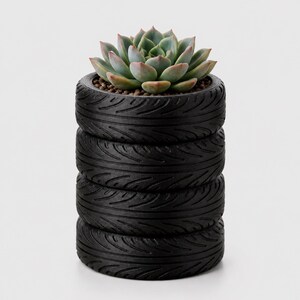 Può includere: Una pianta succulenta con foglie verde chiaro e punte rosa è inserita in un vaso a forma di pila di quattro pneumatici neri. Gli pneumatici presentano dettagliati motivi del battistrada e sono impilati verticalmente in un cilindro.
