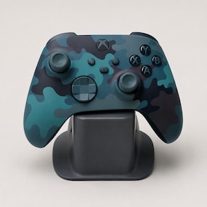 Op de afbeelding: Een draadloze videogamecontroller met een camouflagepatroon in blauwgroen, donkerblauw en zwart. De controller bevat twee analoge sticks, een richtingspad en vier actieknoppen met de letters A, B, X en Y, rustend op een donkergrijze standaard.