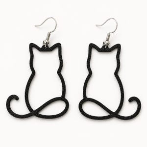 Puede incluir: Un par de pendientes negros con la silueta de un gato. Cada pendiente tiene la forma de un gato sentado con una cola curva y está sujeto a un gancho de metal de tono plateado para colgarlo del lóbulo de la oreja.