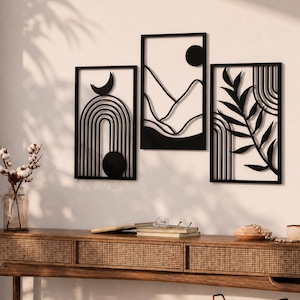 Può includere: Set di tre quadri neri da parete con disegni geometrici e ispirati alla natura. Il set include un pezzo con una luna crescente e un arco, una scena di montagna e un design botanico. L'arte è esposta su una parete bianca sopra una consolle di legno.