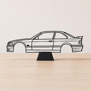 Può includere: Scultura in metallo nero con contorno di auto, montata su una base nera. L'auto presenta un design coupé con spoiler. La scultura è esposta su una superficie di legno marrone chiaro.