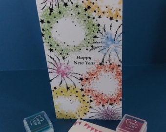 Peut inclure: Une carte de vœux verticale avec un motif de feux d'artifice colorés, des étoiles noires et le texte Happy New Year. Deux encreurs carrés Versa Color cyan et orchidée sont placés à côté d'une carte de visite All Gem's Cards and Crafts.