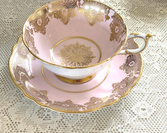 Puede incluir: Un juego de taza y platillo de té rosa con ribetes dorados y diseños florales intrincados. La taza de té tiene un borde festoneado y un asa delicada.