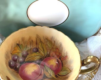 May include: A vintage teacup and saucer set with a gold rim. The teacup features a hand-painted design of peaches, grapes, and leaves on a cream background. The saucer is a light blue colour with a white centre.