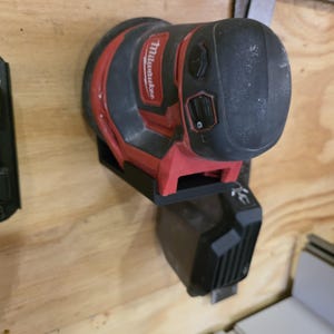 May include: A red and black Milwaukee orbital sander mounted on a wooden surface. The sander is attached to a black base, with a black battery pack below. The sander has a black top with control buttons and the Milwaukee logo.
